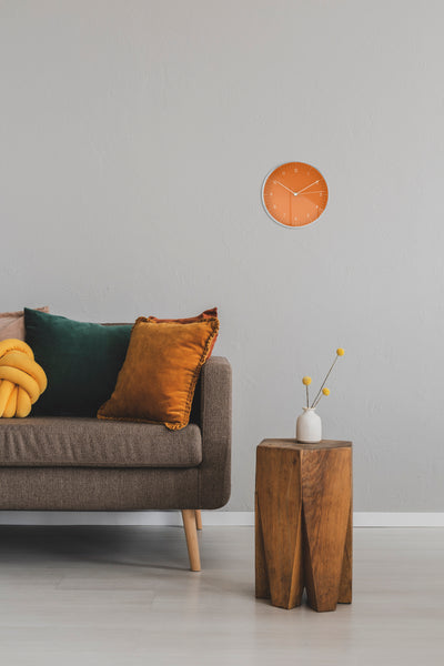 Wanduhr in Orange mit Silbernen Details hängt über grauer Couch mit bunten Kissen und robusten Holz Beistelltisch 