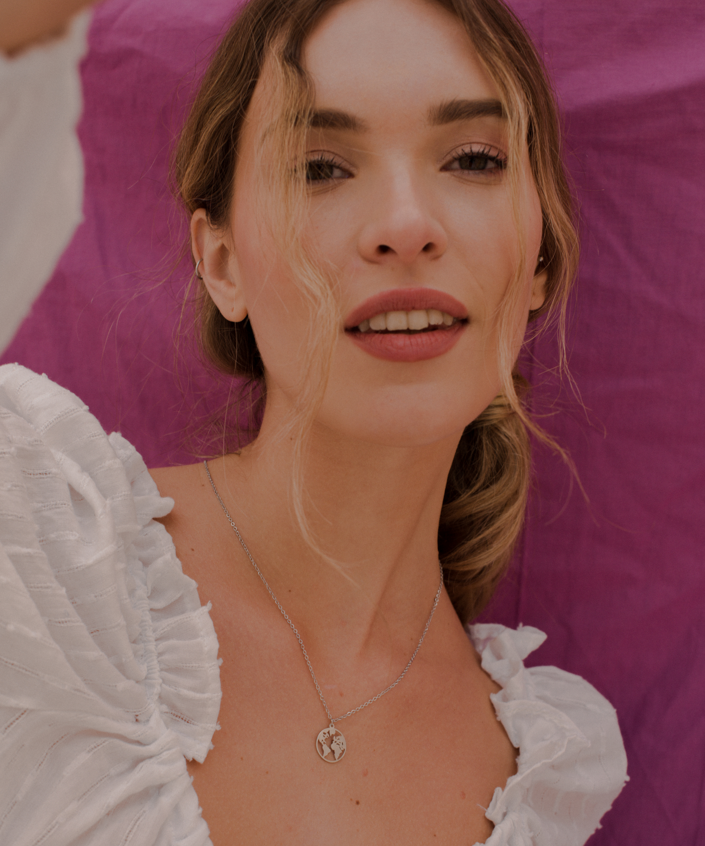 Glückliche Frau trägt feine silberne Kette mit Weltkugel Anhänger im minimalistischen Stil vor pinken Hintergrund