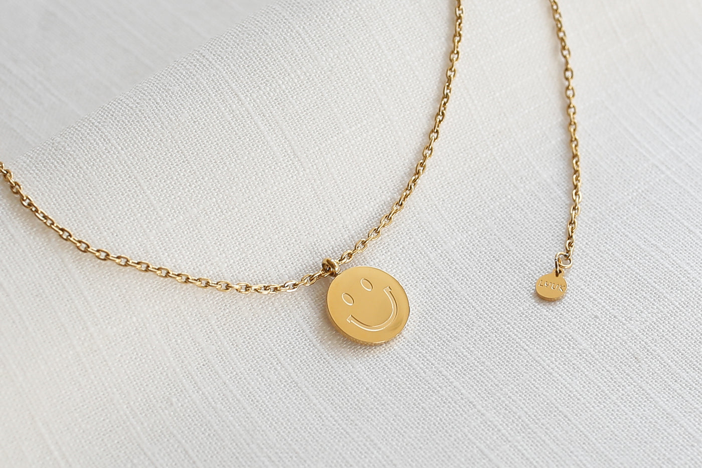 Kette mit Smiley Anhänger und verstellbarer Länge in Gold platziert auf Leinen Stoff.