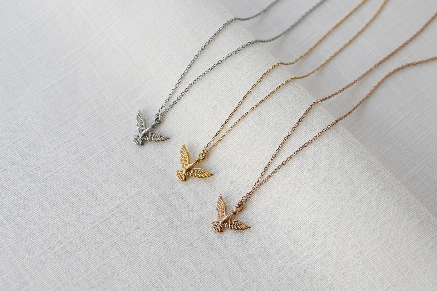 Drei Adler Anhänger Ketten in Silber, Gold und Rosegold ausgebreitet auf Leinen Stoff.