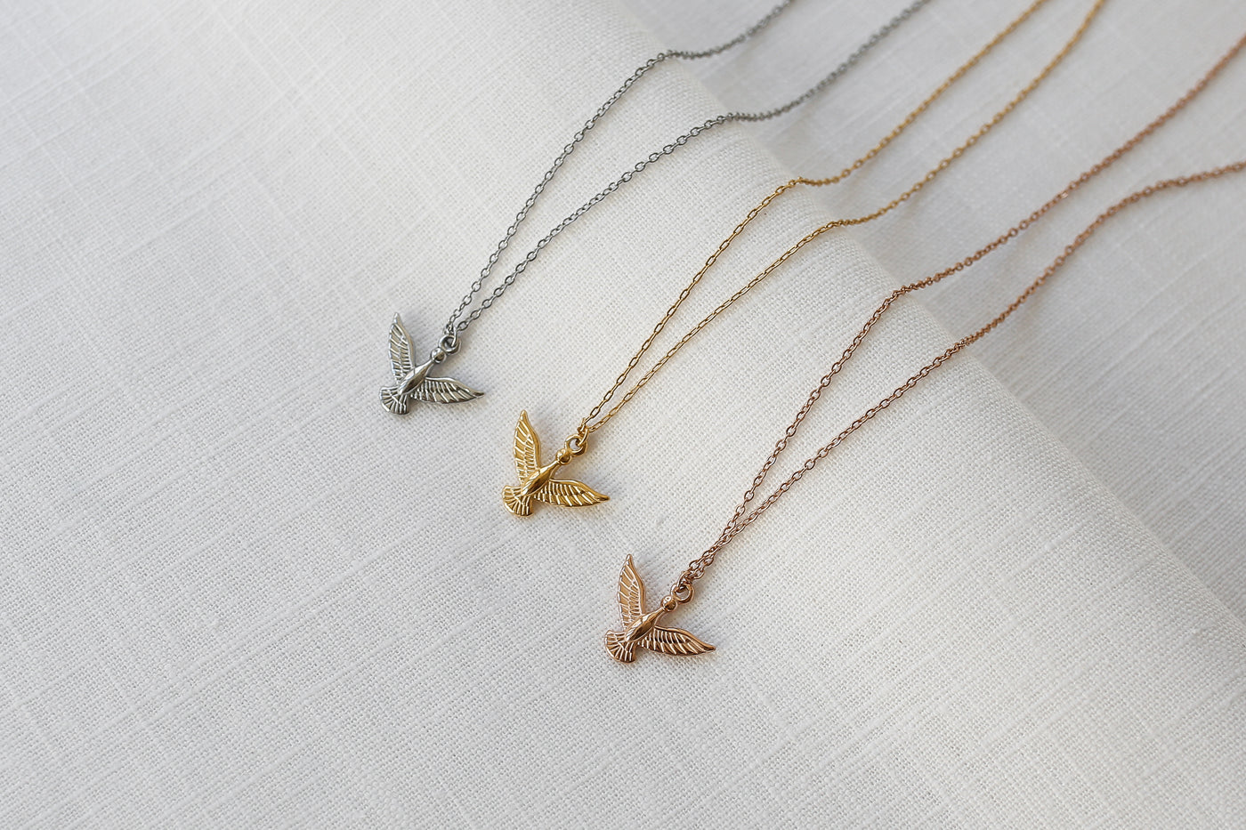 Drei Adler Anhänger Ketten in Silber, Gold und Rosegold ausgebreitet auf Leinen Stoff.