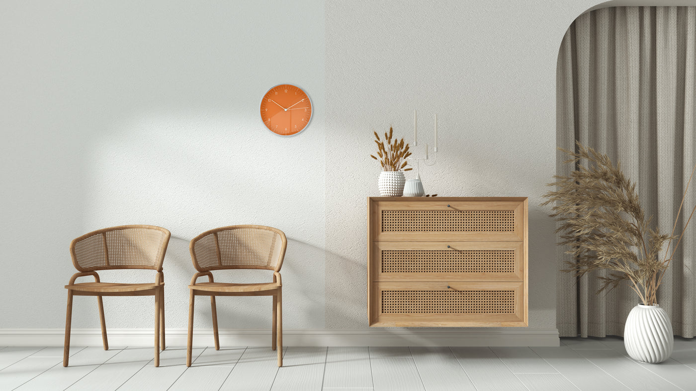 Orangene Wanduhr mit silbernen Akzenten hängt über zwei Holzstühlen und brauner Kommodo 